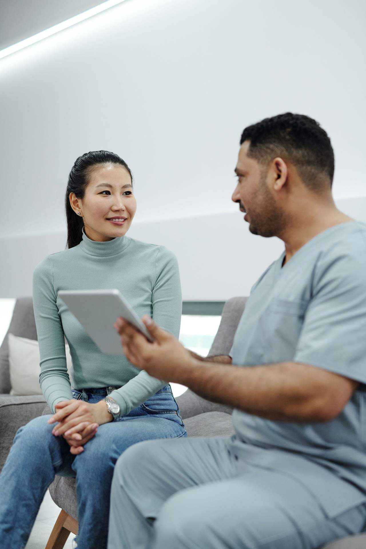 Nha sĩ nam mặc đồng phục xám xanh cầm máy tính bảng giải thích cho một người phụ nữ ngồi cạnh trên ghế sofa ở phòng chờ