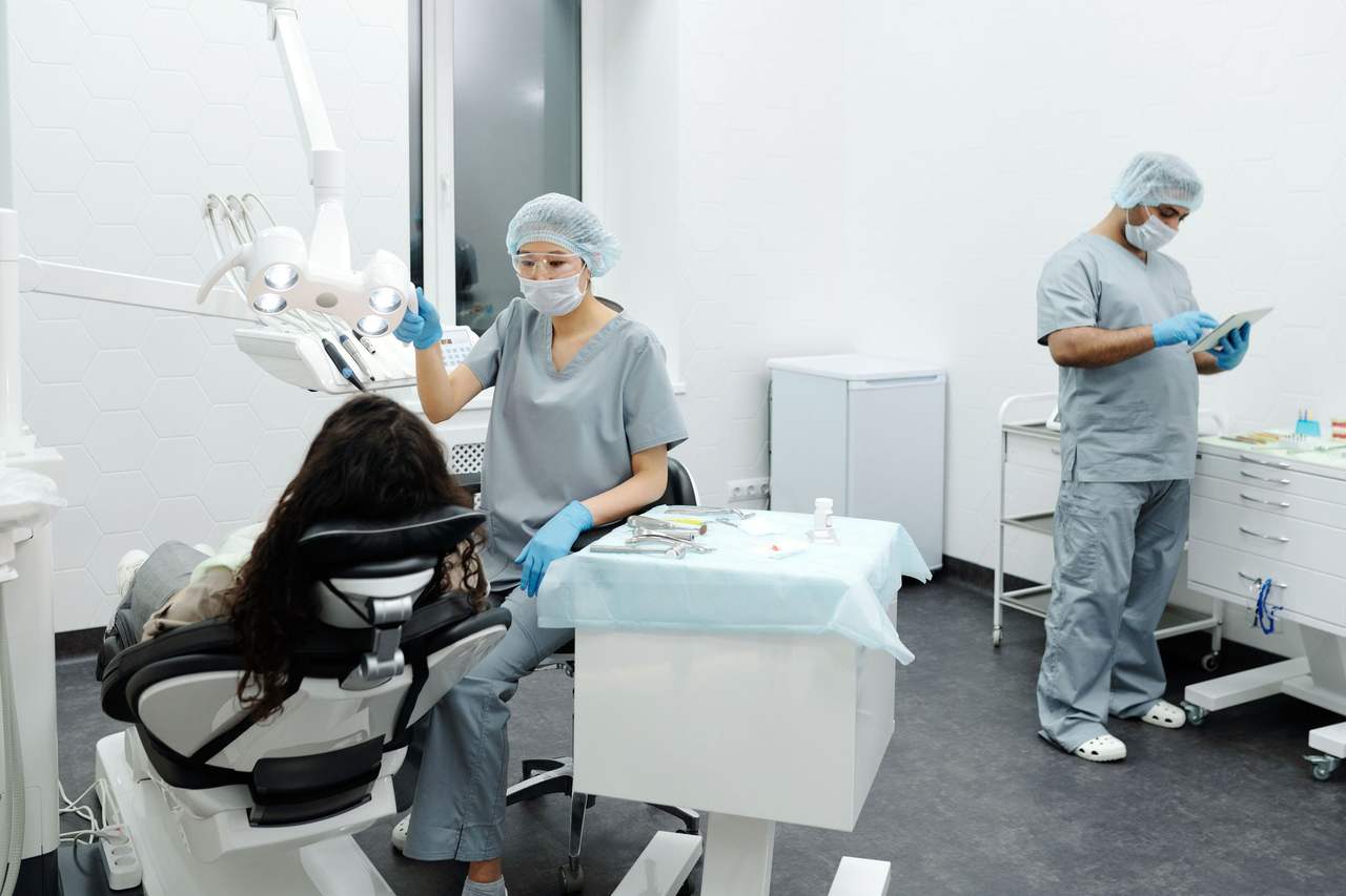 Phòng điều trị sáng màu: nữ nha sĩ chỉnh đèn khám bên người bệnh trên ghế, phía sau một nha sĩ nam xem máy tính bảng cạnh tủ dụng cụ