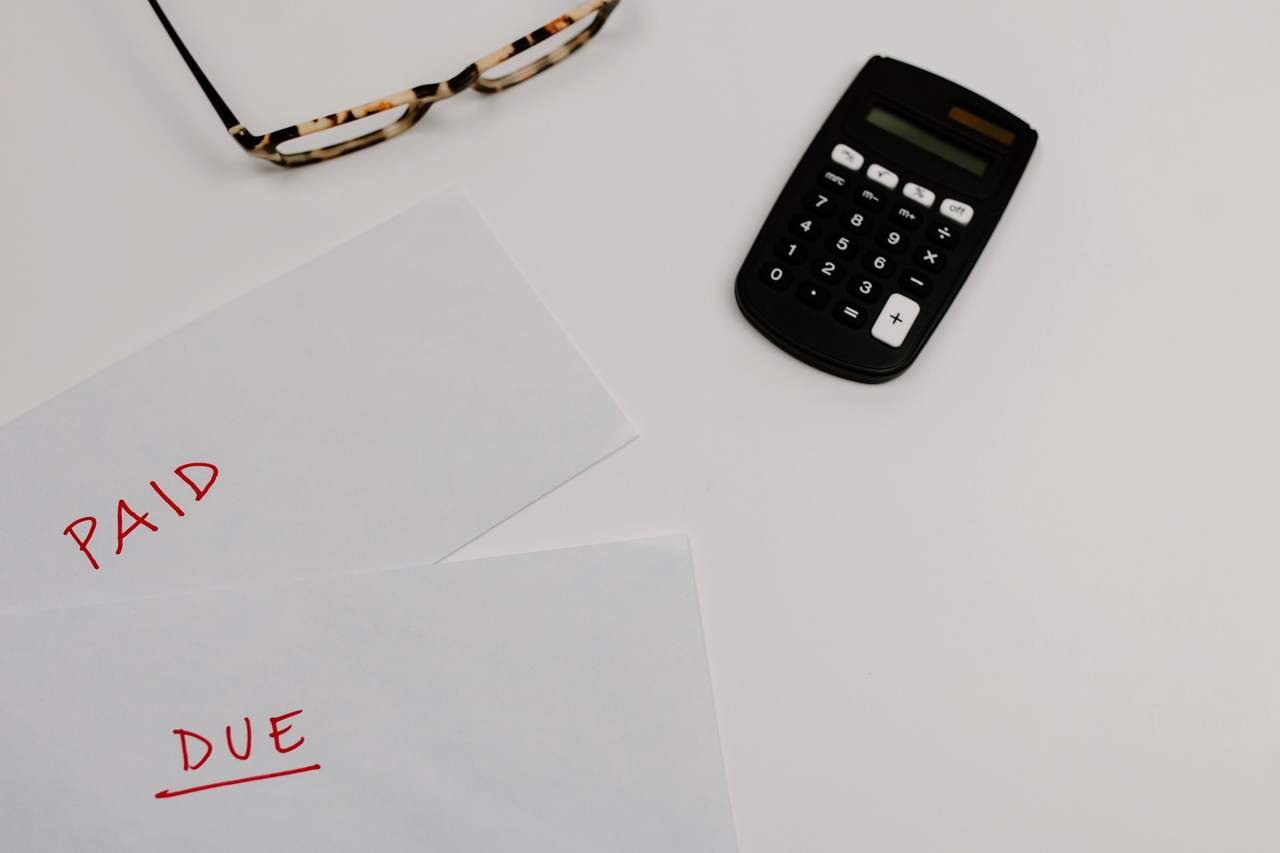 Nhìn từ trên xuống: hai tờ giấy viết tay chữ PAID và DUE màu đỏ, một máy tính cầm tay và cặp kính trên mặt bàn trắng
