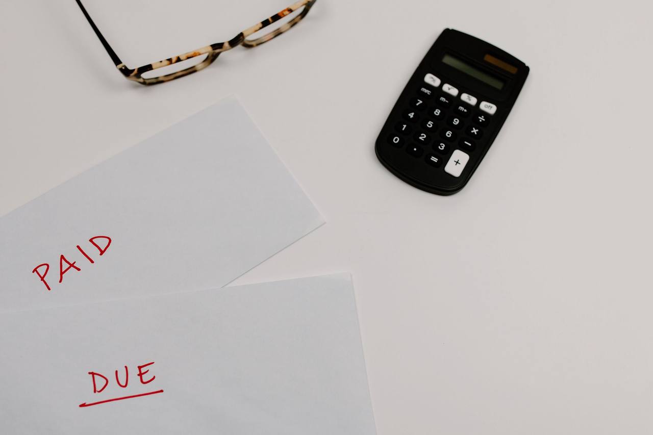 Nhìn từ trên xuống: hai tờ giấy viết tay chữ PAID và DUE màu đỏ, một máy tính cầm tay và cặp kính trên mặt bàn trắng