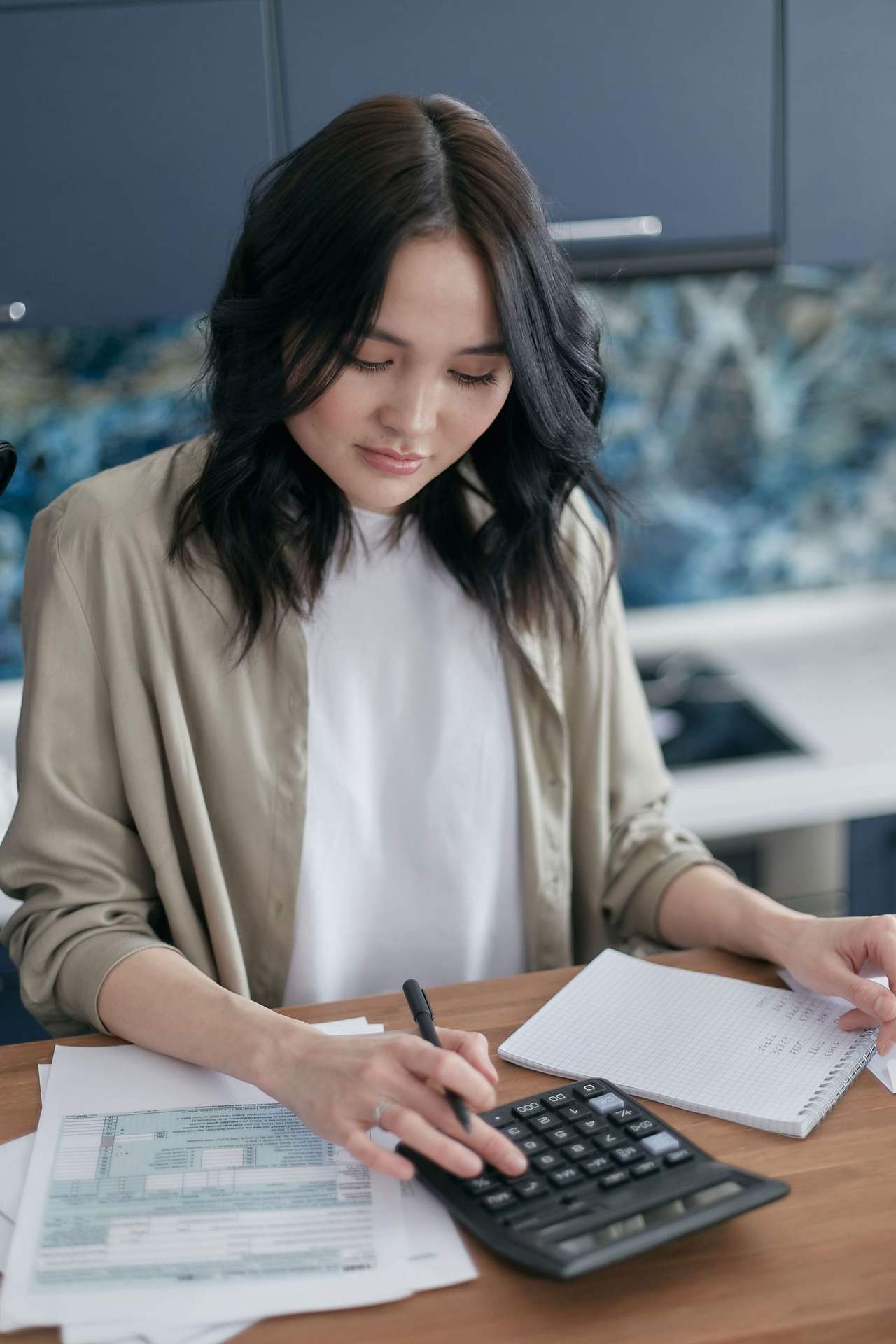 Một người phụ nữ tóc đen ngồi bên bàn gỗ, bấm máy tính cầm tay cạnh xấp giấy tờ và cuốn sổ ghi chép