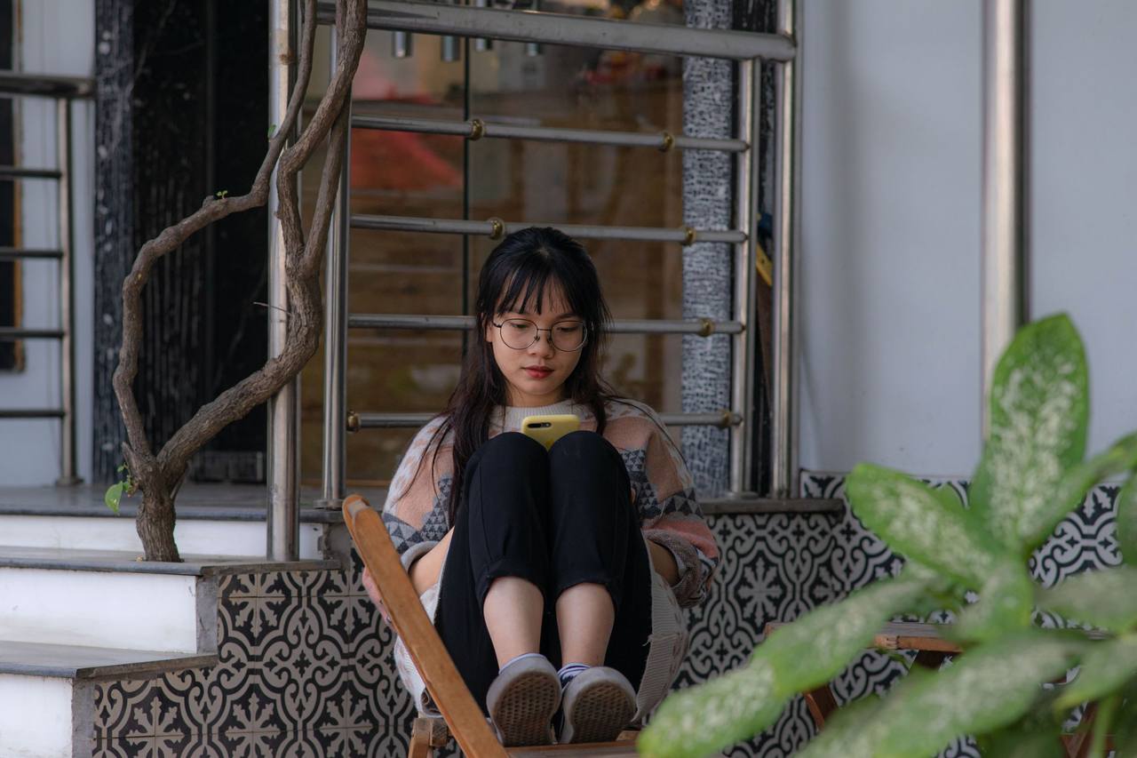 Một cô gái đeo kính, tóc dài, ngồi co chân trên ghế gỗ trước bậc thềm lát gạch bông, chăm chú đọc điện thoại