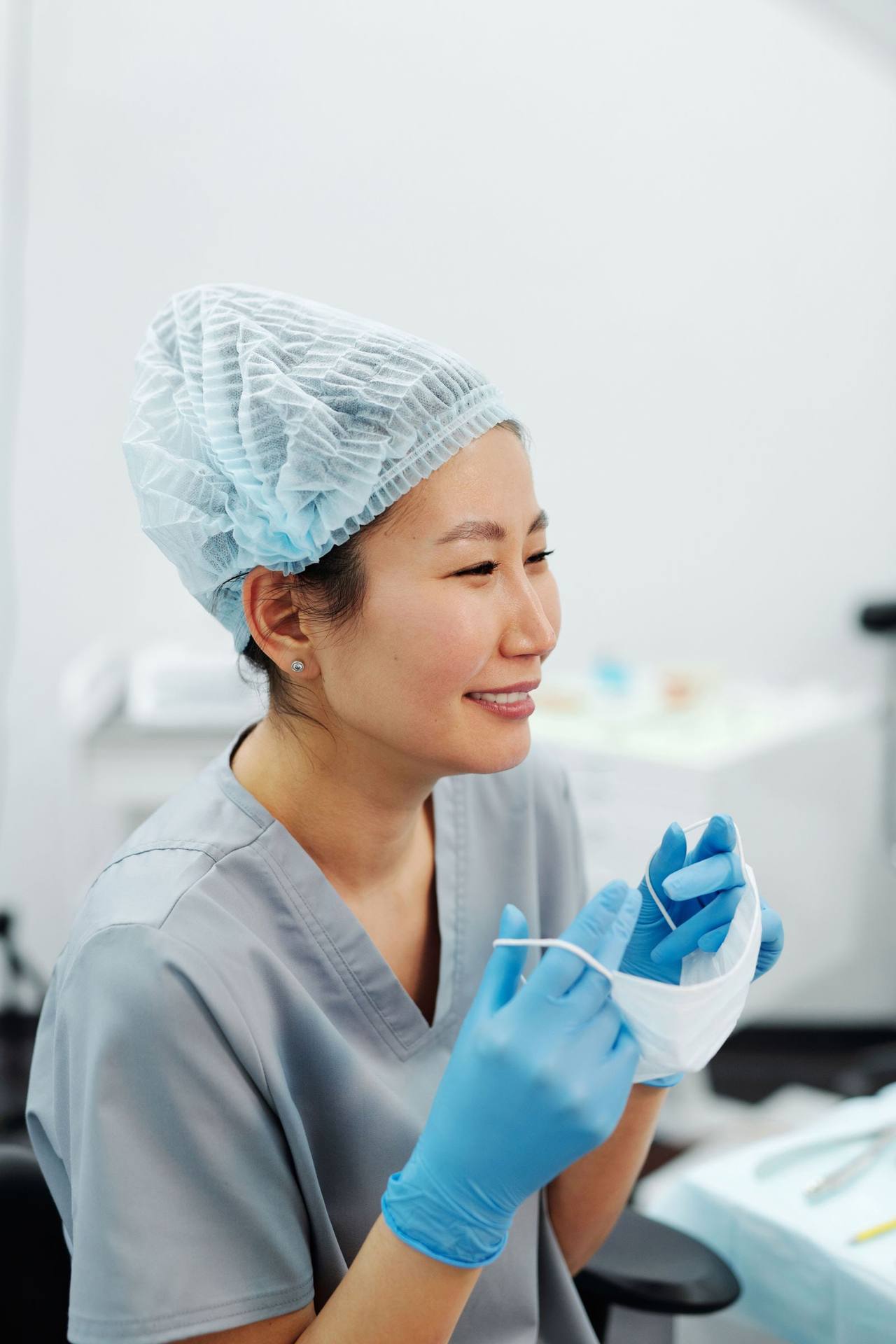Nữ nha sĩ đội mũ trùm tóc, mặc đồng phục xám xanh, đeo găng tay xanh, mỉm cười khi tháo khẩu trang trong phòng điều trị