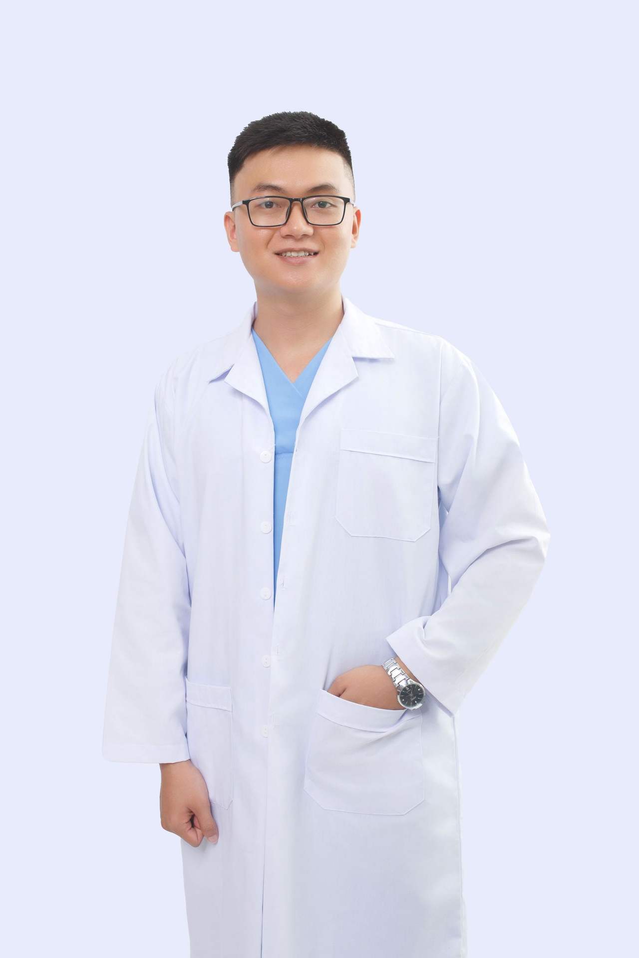 Bác sĩ nam trẻ đeo kính, mặc áo blouse trắng ngoài áo xanh dương, một tay đút túi áo và mỉm cười trên nền xanh xám nhạt