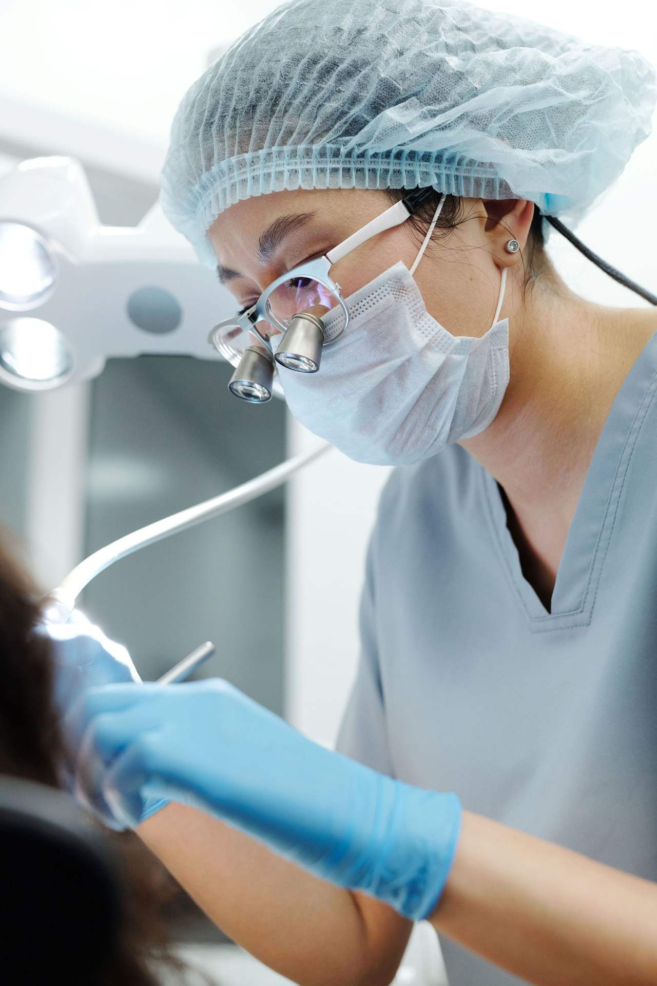 Nữ nha sĩ đội mũ trùm tóc, đeo kính lúp phóng đại và khẩu trang, cầm dụng cụ nha khoa nối dây làm việc cho người bệnh