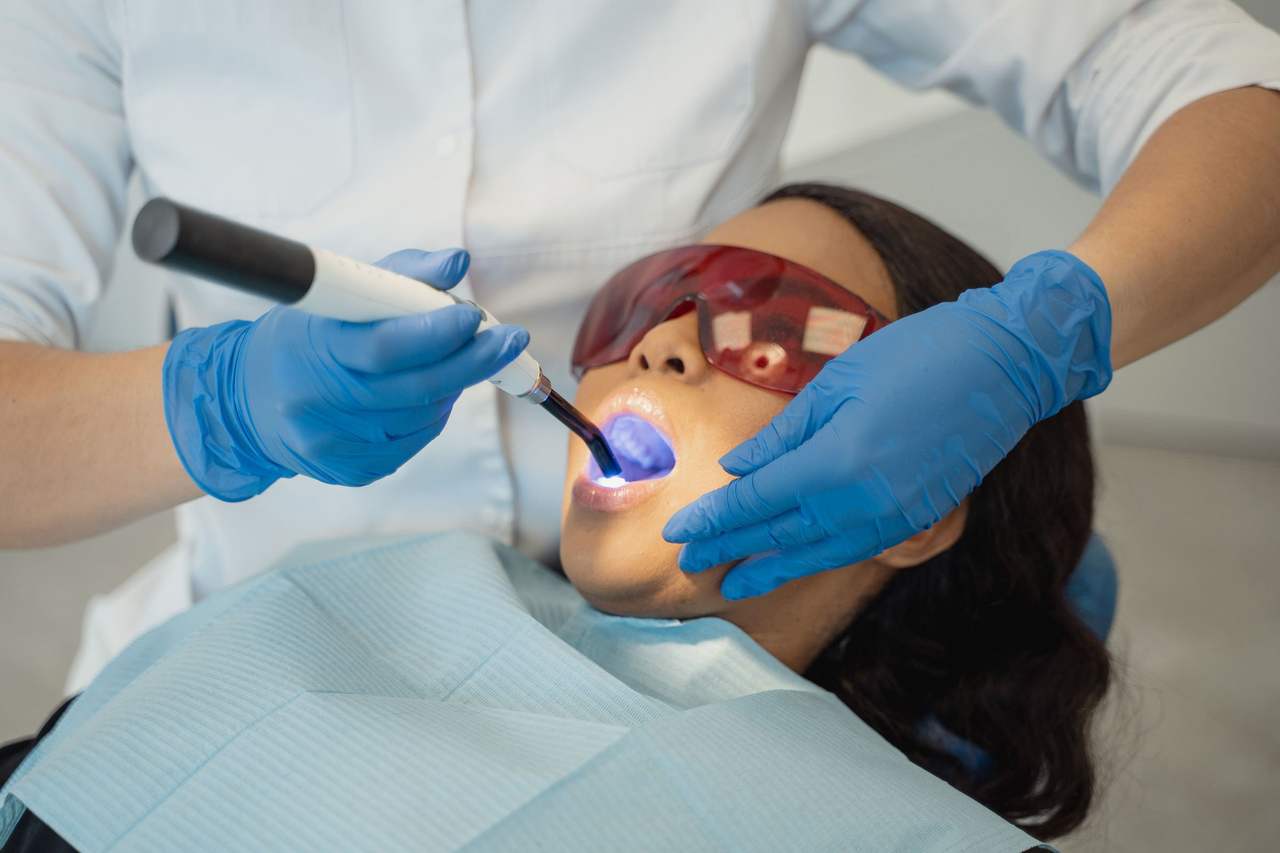 Bác sĩ đeo găng tay xanh chiếu đèn quang trùng hợp vào răng một người bệnh đeo kính bảo hộ màu cam